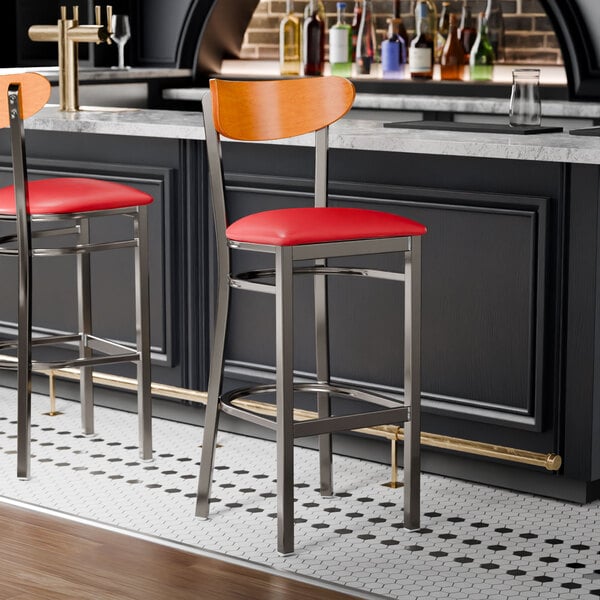 Two Lancaster Table & Seating Boomerang clear coat finish bar stools with red vinyl seats and cherry wood backs at a bar counter.