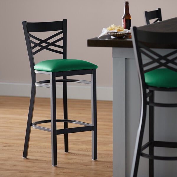 A black Lancaster Table & Seating cross back bar stool with a green vinyl padded seat.