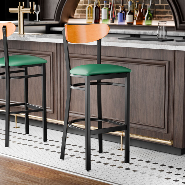 A bar stool with a black metal frame, green vinyl seat, and cherry wood backrest.