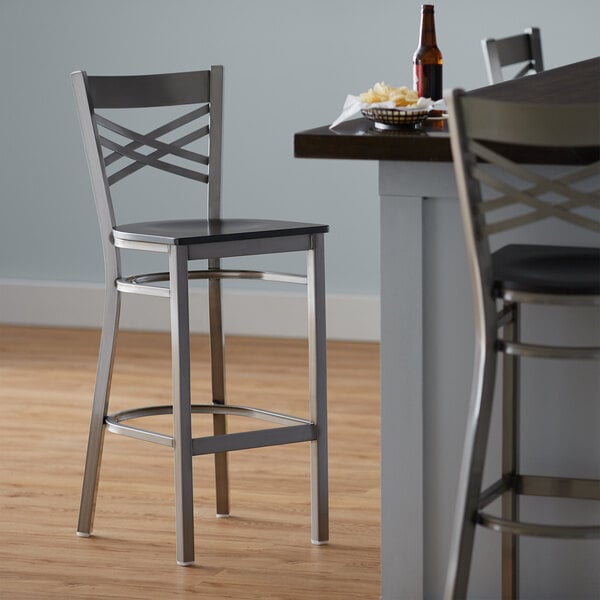 A Lancaster Table & Seating cross back bar stool with a black wood seat next to a table.