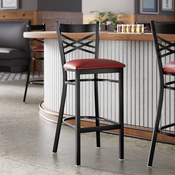 A black metal bar stool with a cross back design and a burgundy vinyl padded seat.