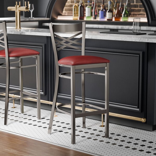 A metal bar stool with a cross back design and a burgundy vinyl padded seat.