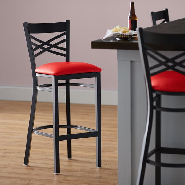 A black Lancaster Table & Seating cross back bar stool with a red vinyl padded seat next to a table.