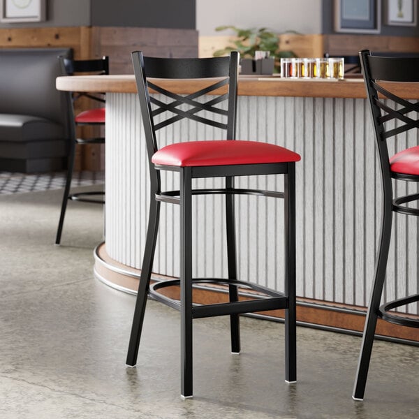 A black metal bar stool with a cross back design and a red vinyl padded seat.