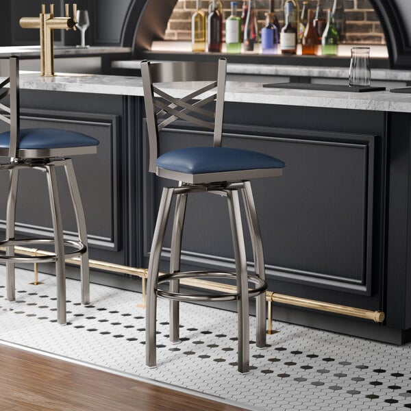 A metal bar stool with a cross back design and a navy blue vinyl padded seat.
