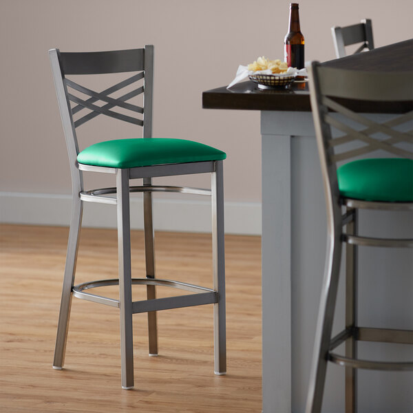 A Lancaster Table & Seating clear coat finish metal cross back bar stool with a green vinyl padded seat.