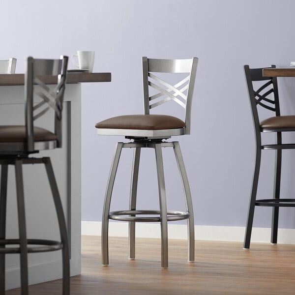 A Lancaster Table & Seating clear coat finish swivel bar stool with a dark brown vinyl padded seat and a chair leg with a cushion.