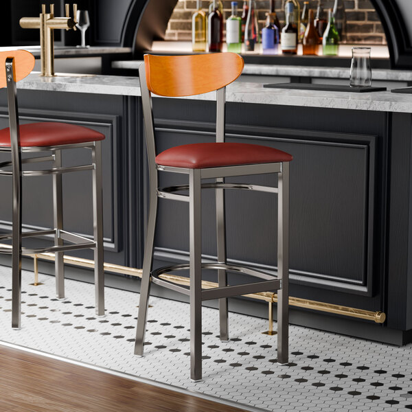 Two Lancaster Table & Seating bar stools with a cherry wood back and burgundy vinyl seats at a bar counter.