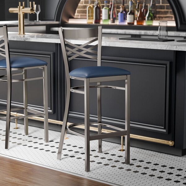 A metal bar stool with a cross back design and a navy blue padded vinyl seat.
