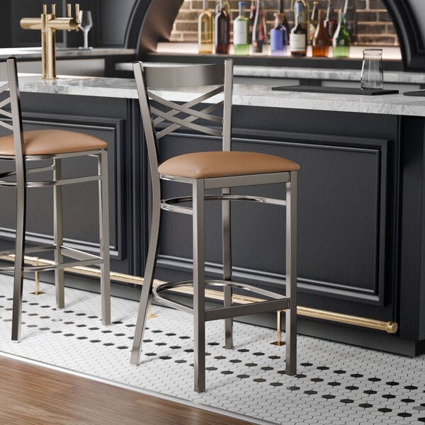A metal bar stool with a cross back design and a light brown vinyl padded seat.