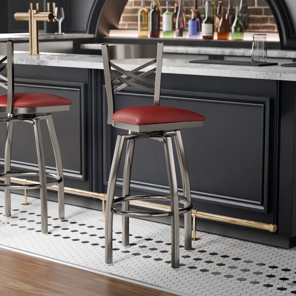 A metal bar stool with a cross back design and a burgundy vinyl padded seat.