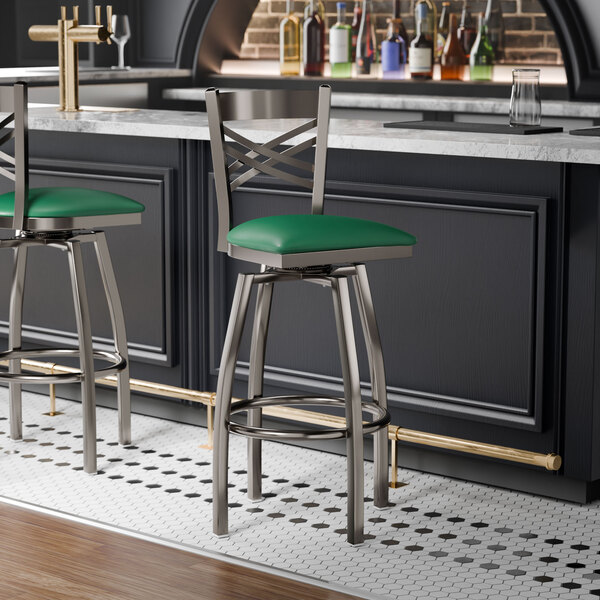 A metal bar stool with a cross back design and a green vinyl padded seat.