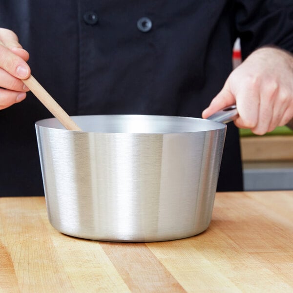 A person stirring a Vollrath stainless steel sauce pan with a wooden spoon.