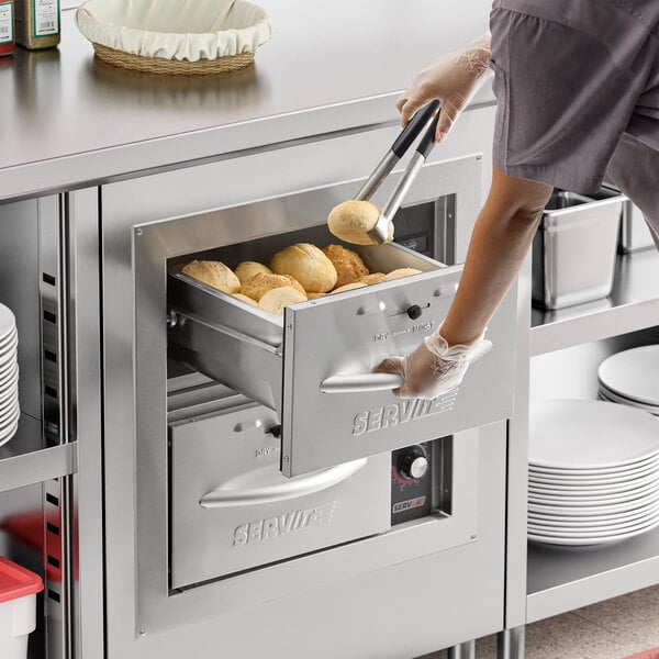 A woman using tongs to put food into a ServIt built-in drawer warmer.