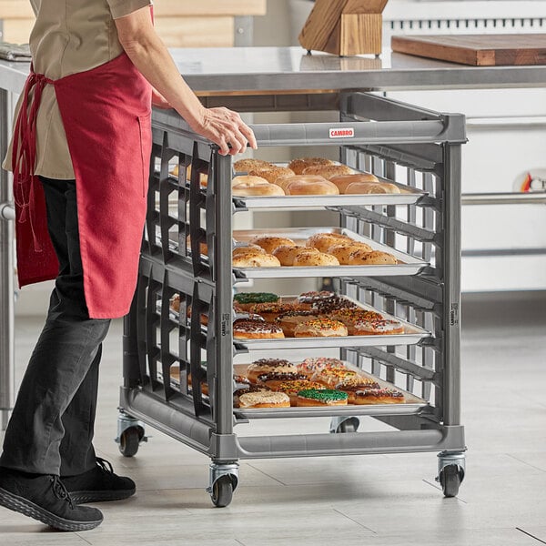 A woman in an apron standing next to a Cambro undercounter sheet pan rack full of food.