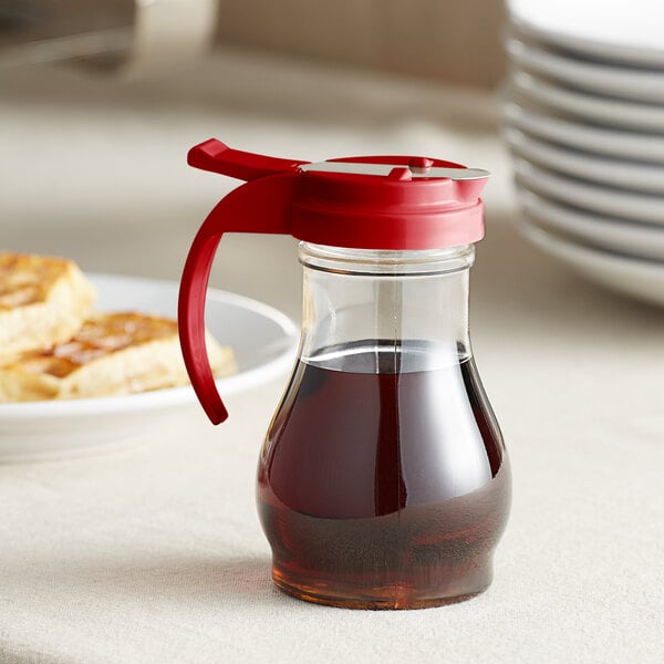 A Vollrath clear polycarbonate syrup server with a red lid on a table with food.