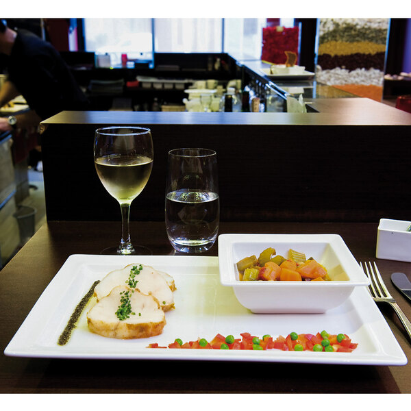 A white square RAK Porcelain bowl with food in it on a table with a plate of food and wine glasses.