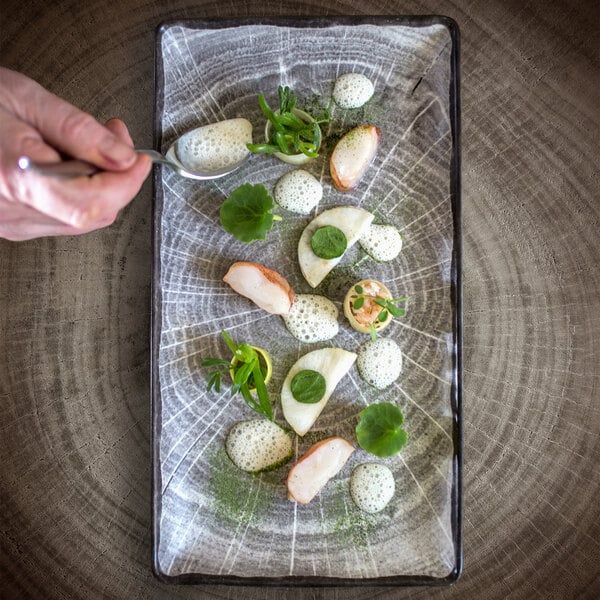 A hand holding a spoon over a RAK Porcelain beech grey rectangular serving platter with food on it.