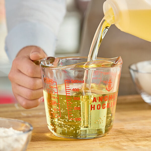 A person pouring liquid into an Anchor Hocking clear glass measuring cup on a kitchen counter.