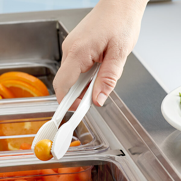 A hand using white plastic tongs to pick up fruit in a container.