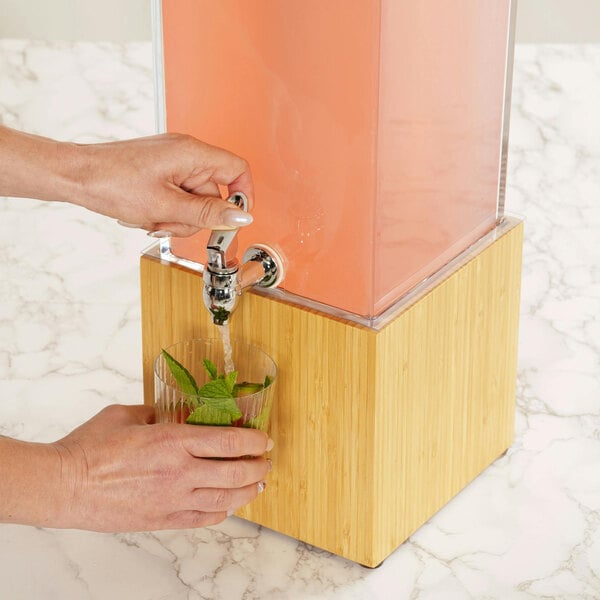 A person filling a glass with water from a Cal-Mil bamboo infusion dispenser.