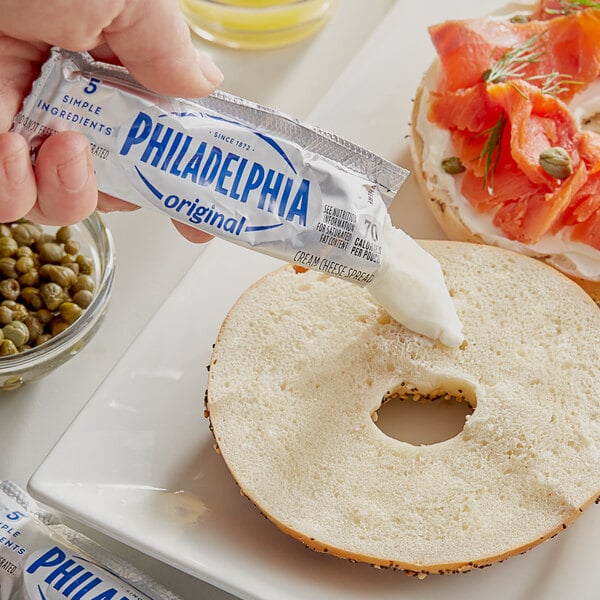 A hand holding a Philadelphia Original Plain Cream Cheese portion packet being spread over a bagel.