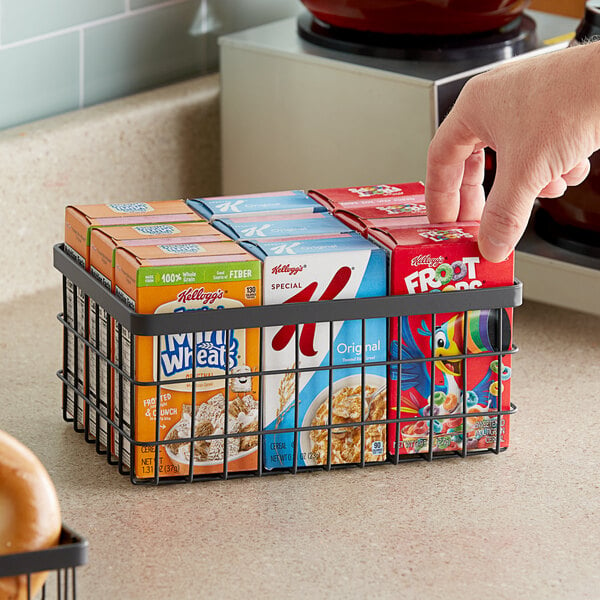 A hand reaching for a box of cereal in a metal gray wire basket.