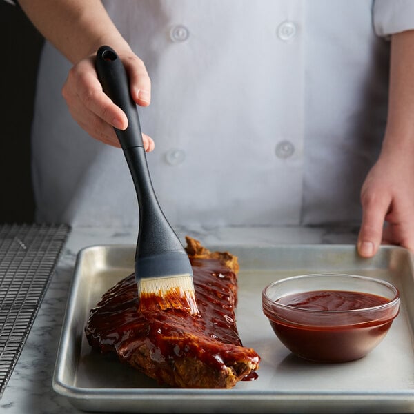 A person using an OXO Good Grips basting brush to spread barbecue sauce on ribs.