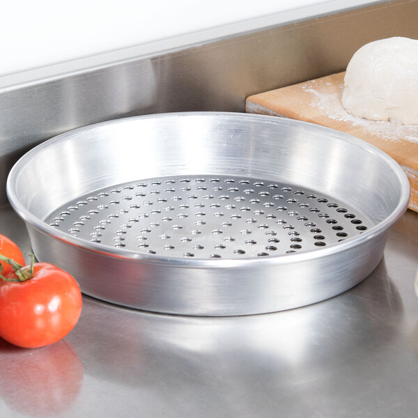 An American Metalcraft heavy weight aluminum pizza pan with holes next to dough and tomatoes.