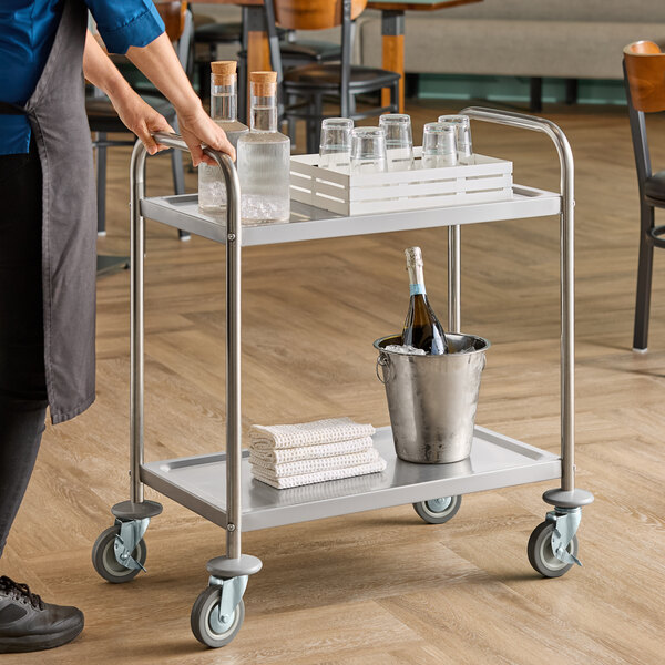 A stainless steel two-shelf utility cart with bottles, glasses, towels, and an ice bucket on its shelves.