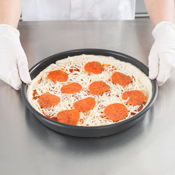 A person holding a Chicago Metallic deep dish pizza with cheese and pepperoni on a pan.