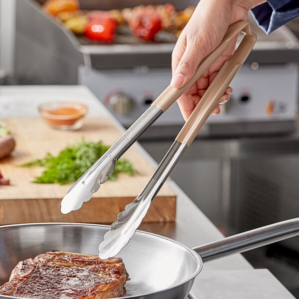 A person using Vollrath stainless steel tongs with tan Kool-Touch handles to cook steak.