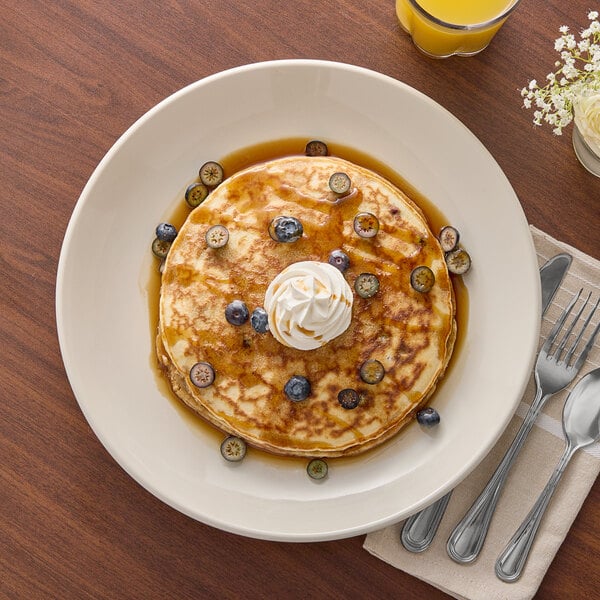 An Acopa round ivory stoneware plate with pancakes topped with whipped cream and blueberries on a table with a spoon and fork.