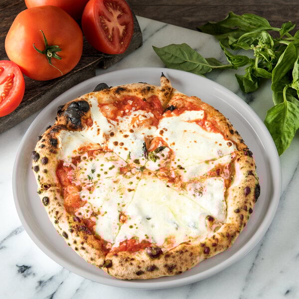 A Carlisle Griege melamine dinner plate with a pizza on it next to tomatoes and basil.