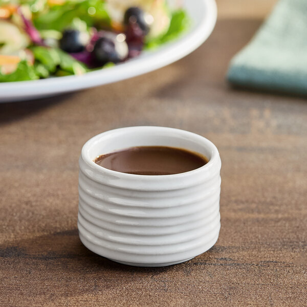A white American Metalcraft porcelain sauce cup filled with brown sauce on a table next to a plate of food.