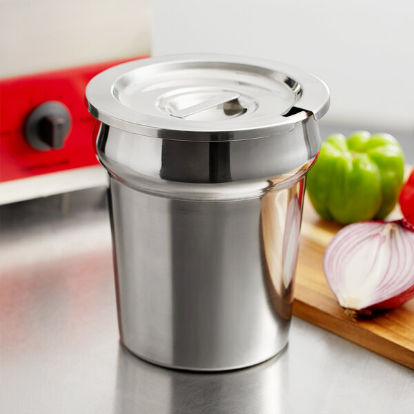 A silver stainless steel vegetable inset with a notched lid on a cutting board next to green bell peppers and onions.