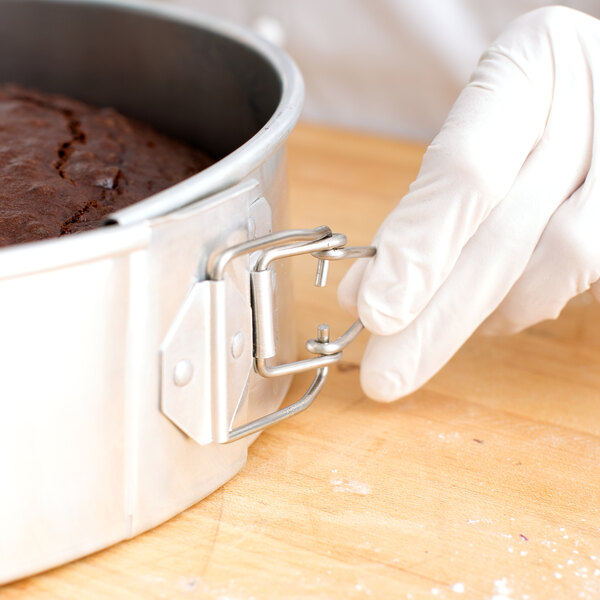 A person in gloves holding a Chicago Metallic aluminum springform cake pan with a brown cake inside.