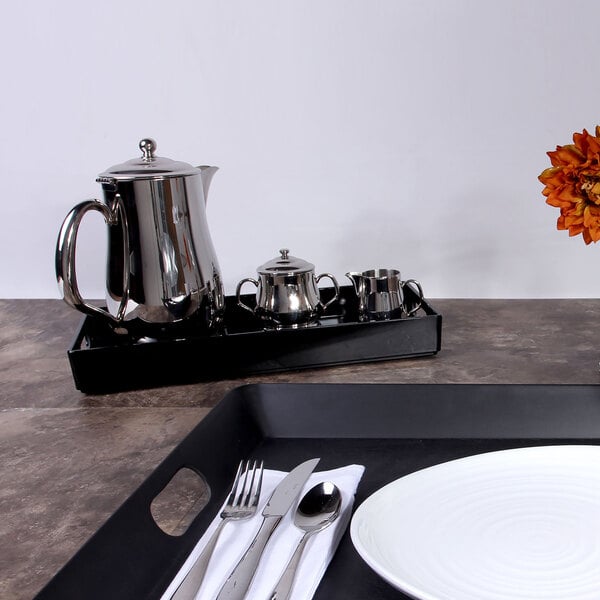 A black rectangular melamine room service tray with silverware and a teapot on it.