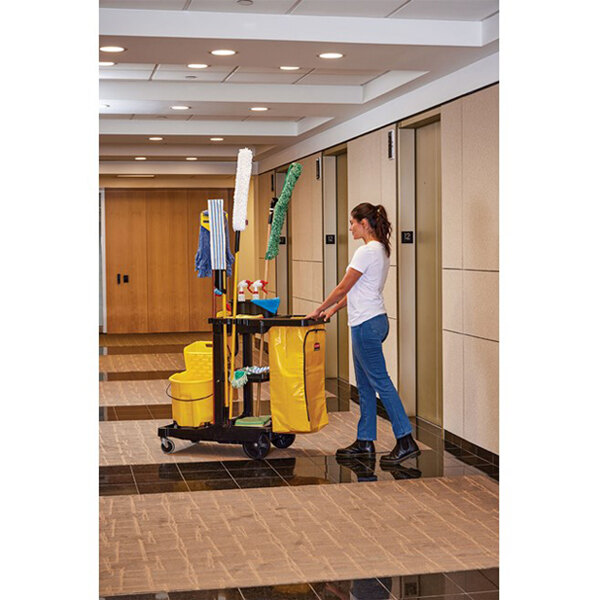 A janitorial cart with cleaning supplies and a yellow bag being pushed by a person in a hallway.