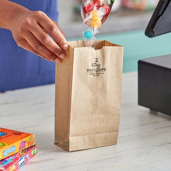 A hand holding a Duro brown paper bag filled with candy in front of a cash register.