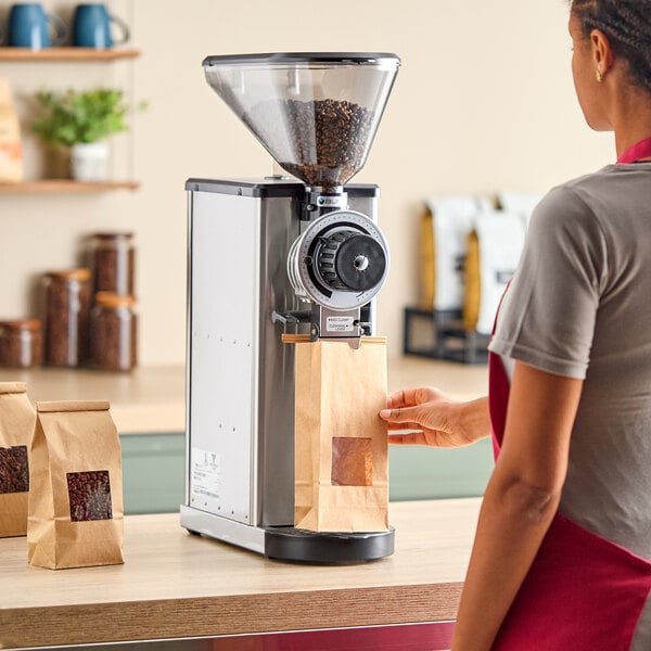 A woman in a red apron using a Bunn stainless steel coffee grinder to grind coffee beans on a counter.