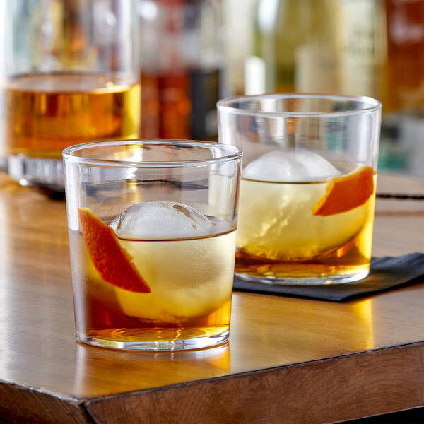 Two Arcoroc old fashioned glasses of whiskey with an orange slice on a table.