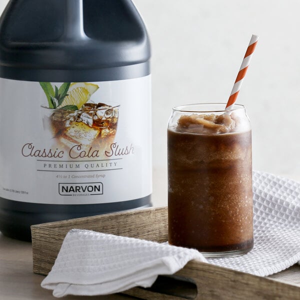 A bottle of Narvon Cola Slushy Syrup next to a glass of brown liquid with a straw.