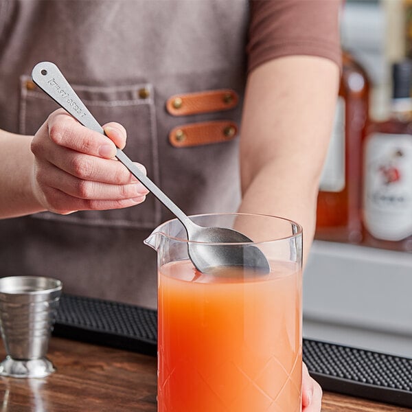 A person using a Barfly stainless steel measuring spoon to mix a drink in a glass of orange liquid.