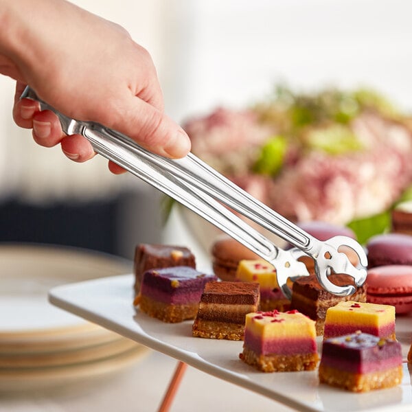 A hand using Acopa stainless steel tongs to pick up a pink macaroon from a plate of desserts.