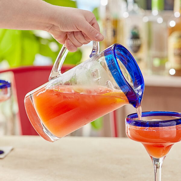 A person pouring a glass pitcher of liquid into a glass with a blue rim.