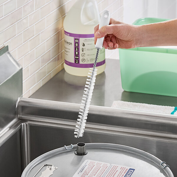 A hand holding a Carlisle white polyester cleaning brush over a metal container.
