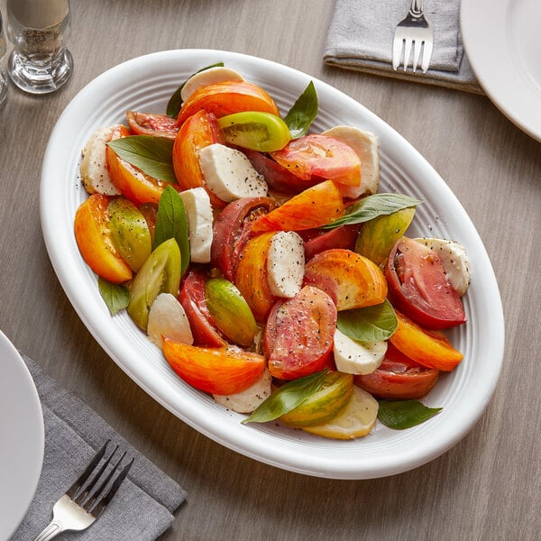 A Tuxton white oval china coupe platter with tomatoes and basil slices on a table.