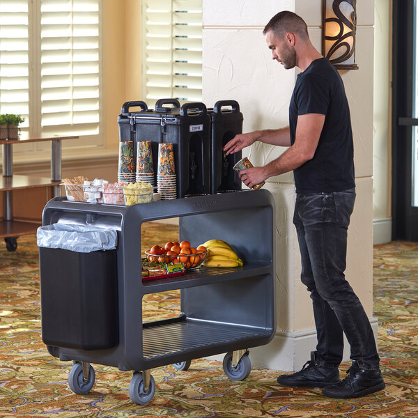 A man standing next to a Cambro charcoal gray service cart with food on it.