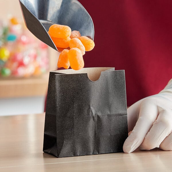 A gloved hand using a spoon to put orange candy in a black American Metalcraft snack bag.